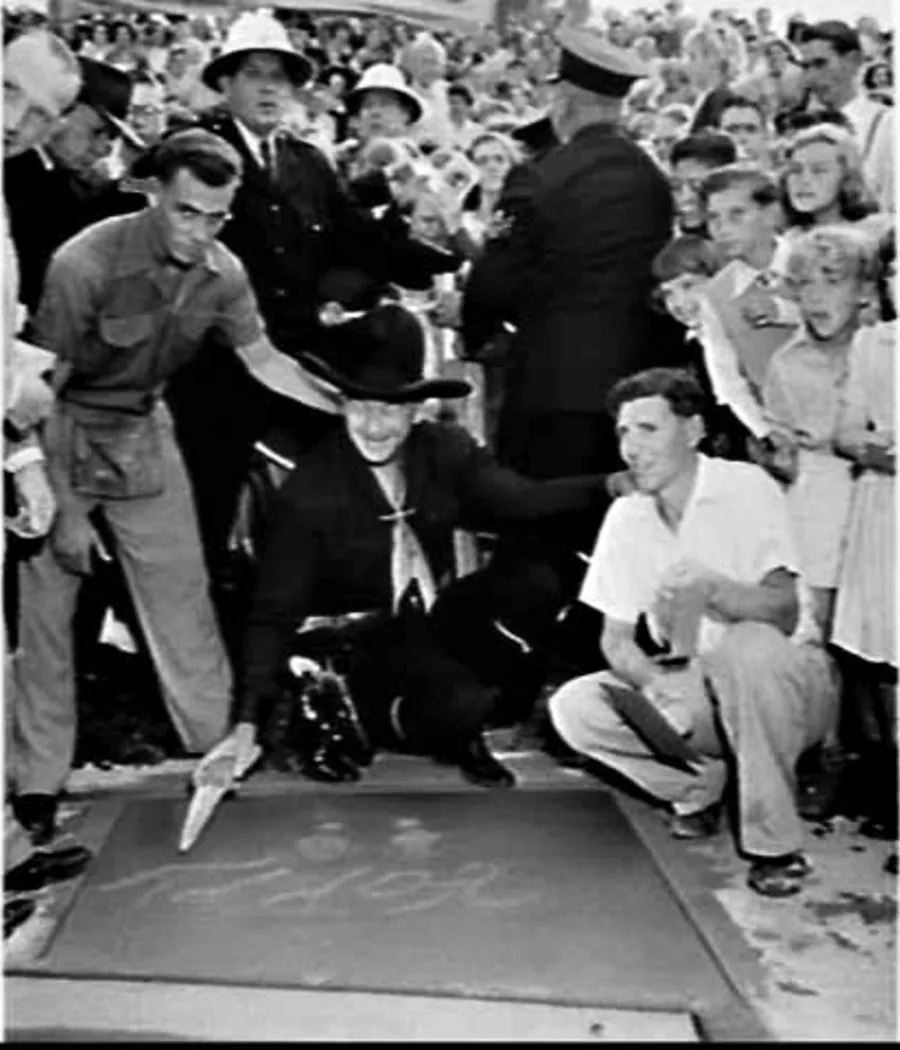 Boyd in his black hat crouches by a slab of wet cement and writes in it with a trowel, between a workman and a young man; police and children crowd behind.