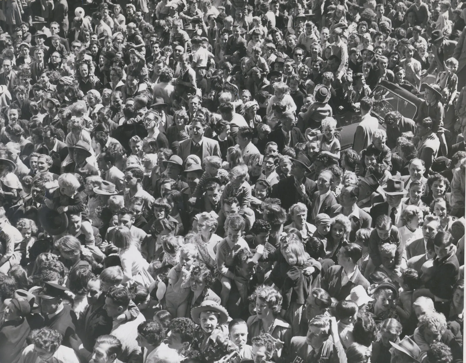 Looking down on a packed street crowd, hundreds of faces turned up: children on their fathers’ shoulders, small boys in cowboy hats, women and men in their Saturday hats, a car swamped at the right.