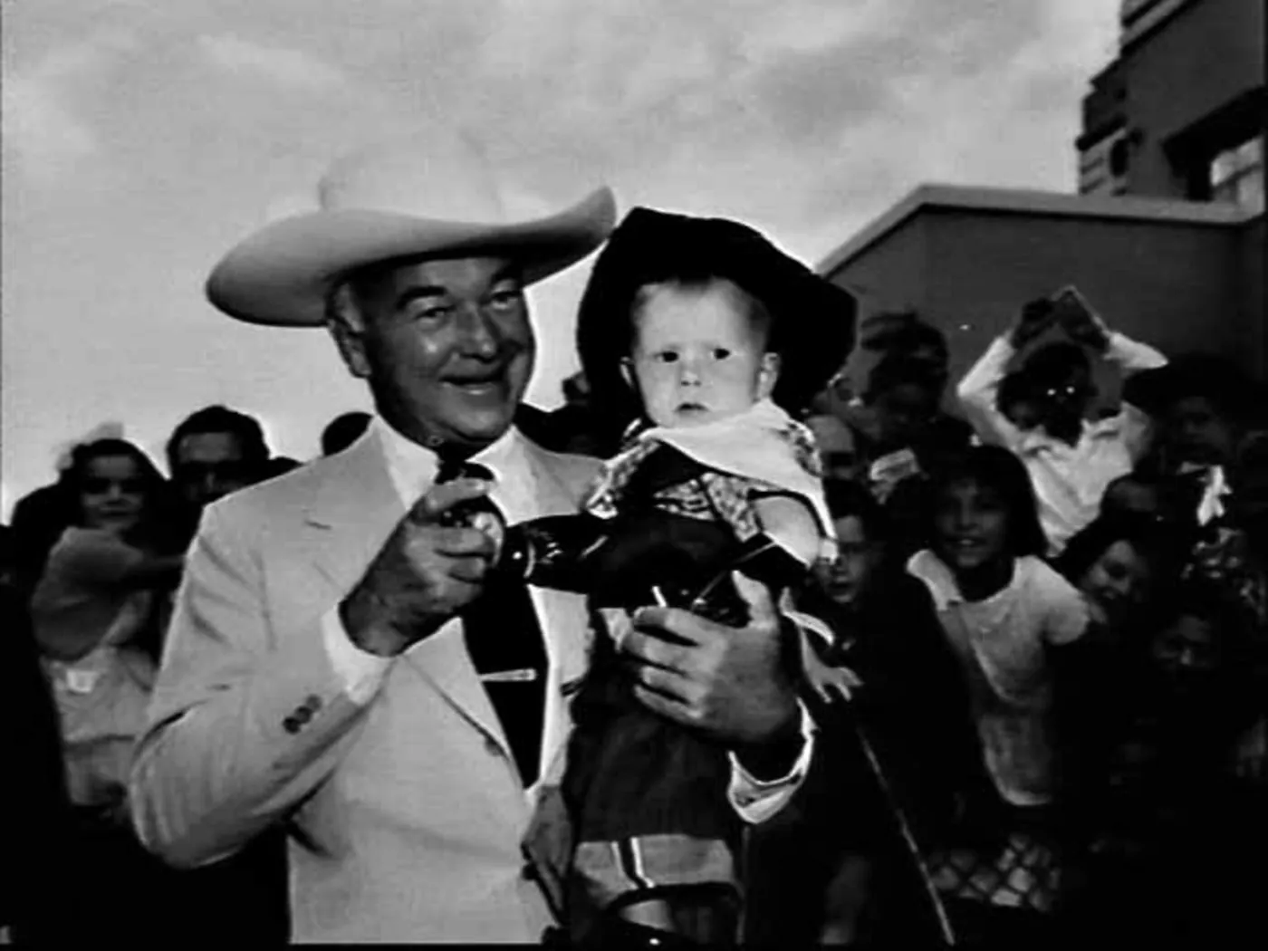 Boyd in a white hat and pale suit, smiling, holds a solemn toddler in an oversized black cowboy hat and neckerchief who grips a toy pistol; children and women crowd behind.