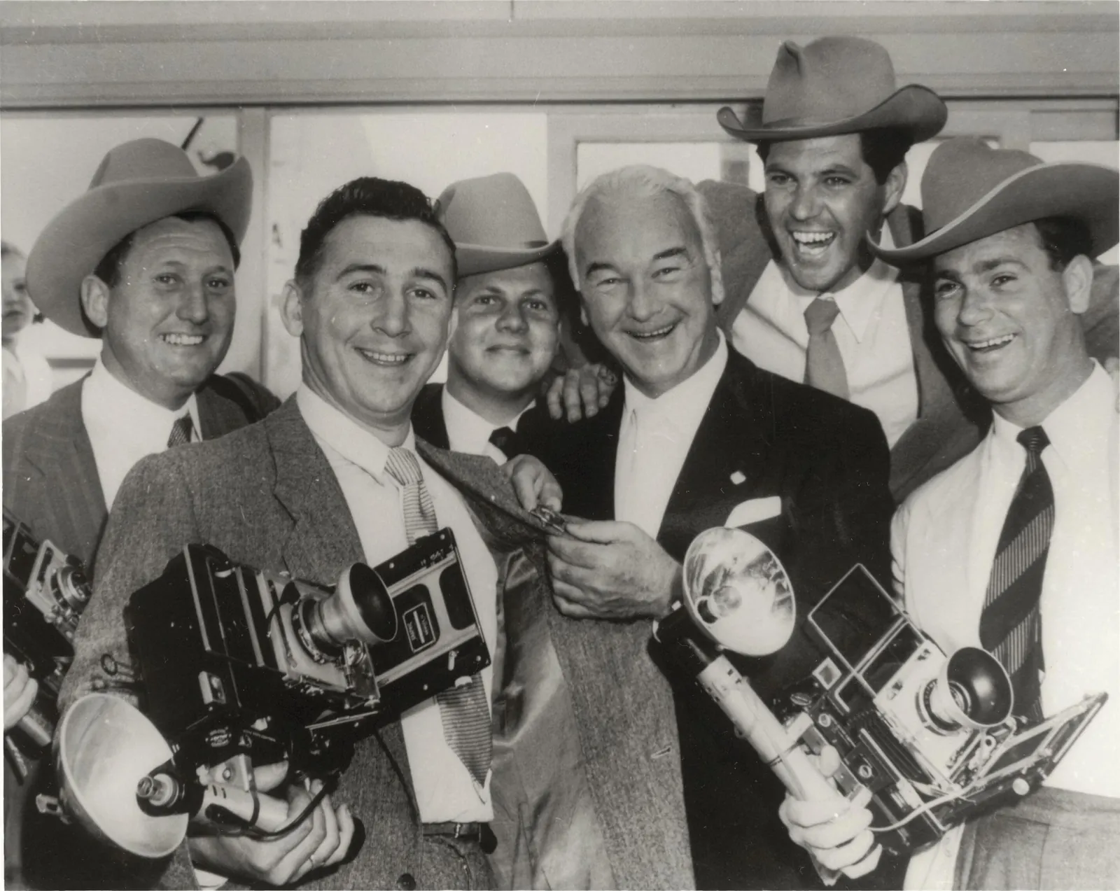 Six men crowded together and grinning: Boyd in the middle, bareheaded and white-haired, tugging the lapel of the man beside him, among five photographers in cowboy hats holding their flash cameras.