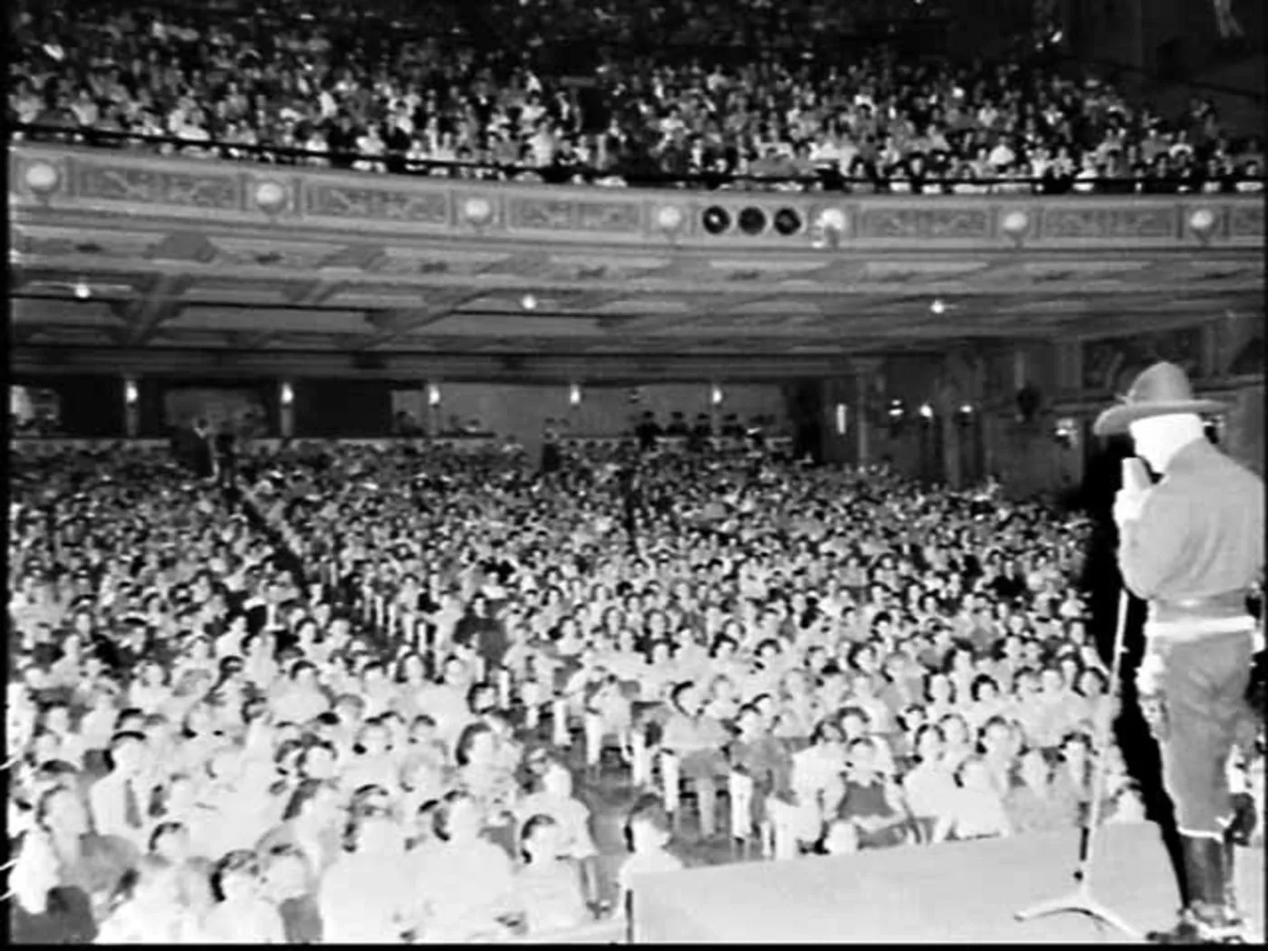 From the stage of a great picture palace: the stalls and the dress circle full of children to the back wall, and at the right edge of the stage a man in a broad-brimmed hat at a microphone.